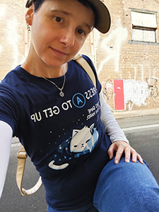 A white non-binary person in a blue tee shirt with a cat on it, and new jeans, posing in front of a brick wall covered with grafitti