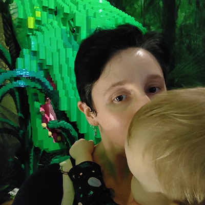 Miriam's face is obscured by the head of the blond baby that ne is kissing. There's a lego dinosaur eating grass in the background.