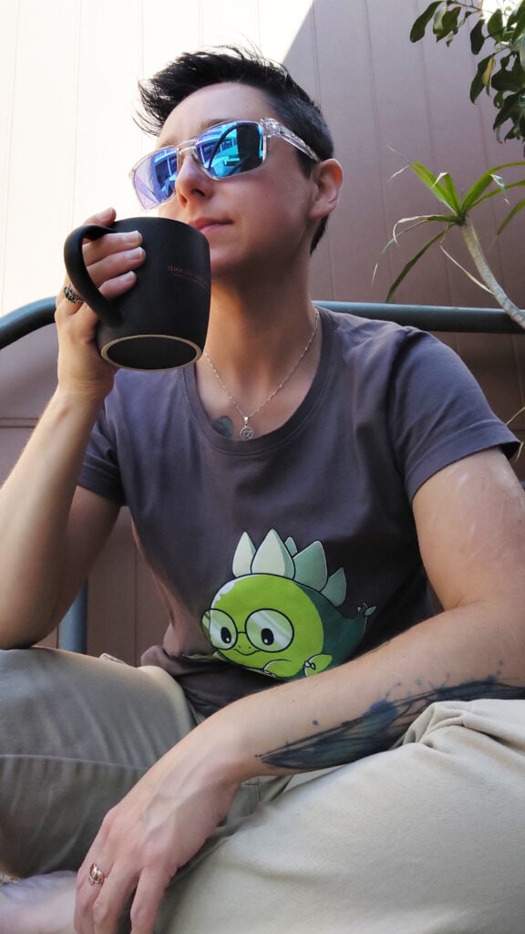 A pale-skinned enby sits on their back step drinking coffee. They're average sized, tattooed, and have short black hair. They're wearing sand coloured cargo pants and a dark grey tee shirt with a dinosaur on it - a thesaurus.